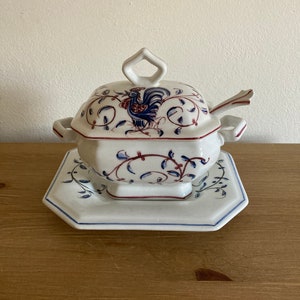 May include: White porcelain gravy boat with a rooster design and a matching lid and serving spoon. The gravy boat has a square base and a rounded top. The rooster is painted in blue, red, and black. The gravy boat is on a white porcelain plate with a matching design.