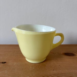 May include: A vintage yellow glass mixing bowl with a handle and a pouring spout. The bowl has a rounded shape and a white interior. The bowl is sitting on a wooden surface.