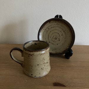 May include: A speckled brown and beige ceramic coffee cup and saucer set. The cup has a rounded handle and the saucer has a circular design.