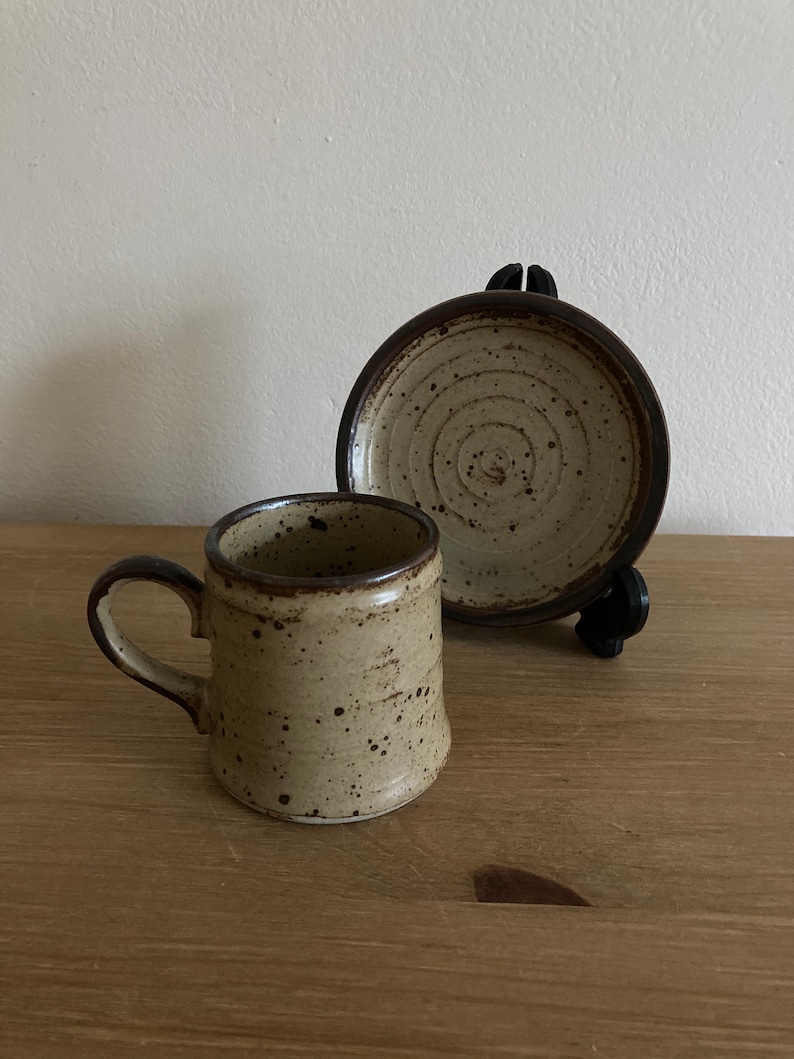 May include: A speckled brown and beige ceramic coffee cup and saucer set. The cup has a rounded handle and the saucer has a circular design.
