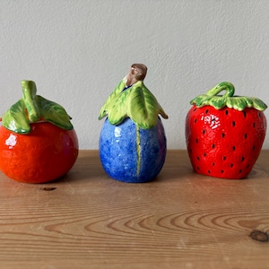 May include: Three ceramic fruit-shaped containers with lids. The first is a round orange with a green stem and leaves. The second is a blue plum with a brown stem and green leaves. The third is a red strawberry with green leaves on top.