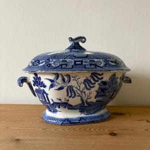 May include: A blue and white ceramic serving bowl with a lid. The bowl features a classic Willow pattern design with handles on each side. The lid has a decorative handle. The bowl is on a wooden surface.