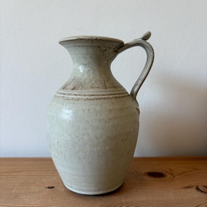 May include: A ceramic pitcher with a speckled off-white glaze and a wide, flared rim. The vessel features a rounded body, a narrow neck, and a single curved handle attached to the side. Subtle brown horizontal bands accent the neck area.
