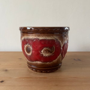 May include: A brown and red ceramic planter with a floral design. The planter has a rounded bottom and a slightly flared top.