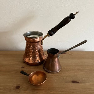 May include: Three copper coffee pots with wooden handles. The largest pot has a hammered finish. The smallest pot has a small ladle.