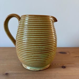 May include: A handmade ceramic jug with a rounded body and a curved handle. The jug is light green with horizontal brown stripes. The spout is on the top, and the jug sits on a small base. The jug is on a wooden surface.