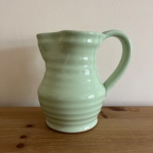May include: A light green ceramic jug with a rounded body and a slightly flared lip. The jug has a textured, ribbed design and a handle.