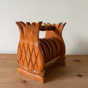 May include: A set of wooden coasters and a holder, all in a warm orange hue. The holder is shaped like a pineapple, with a handle and diamond carvings. The coasters are round and stacked inside. The holder's dimensions are approximately 20 cm.
