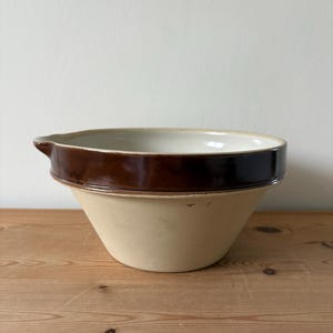 May include: A vintage ceramic mixing bowl in beige and brown. The bowl has a pouring spout and a wide, rounded shape. It features a dark brown band around the rim and a lighter beige base. The interior is white.