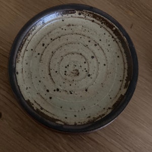 May include: A small, round, ceramic plate with a brown rim and a white center. The plate has a concentric circle design and brown speckles throughout.