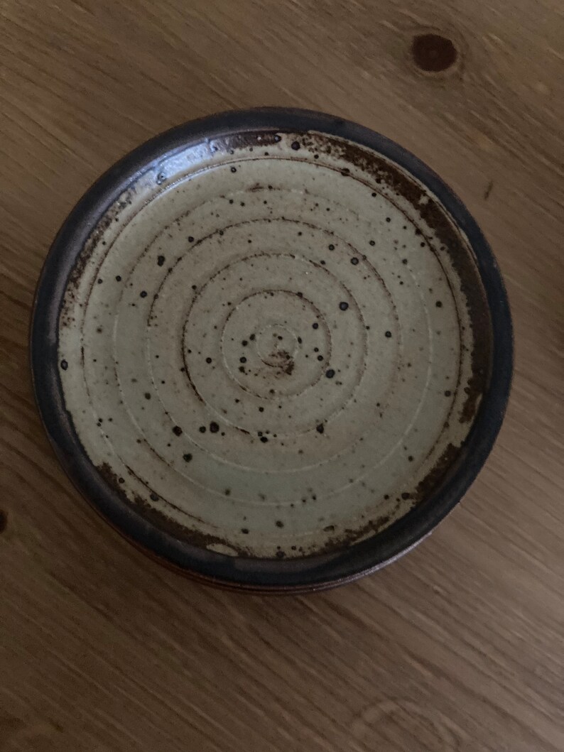May include: A small, round, ceramic plate with a brown rim and a white center. The plate has a concentric circle design and brown speckles throughout.