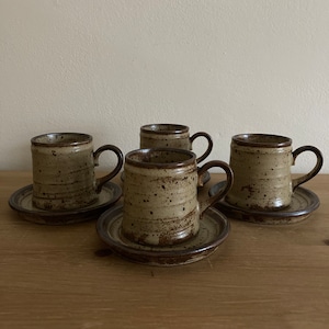 May include: Set of four brown and beige ceramic coffee mugs with matching saucers. The mugs have a speckled glaze and a brown rim.