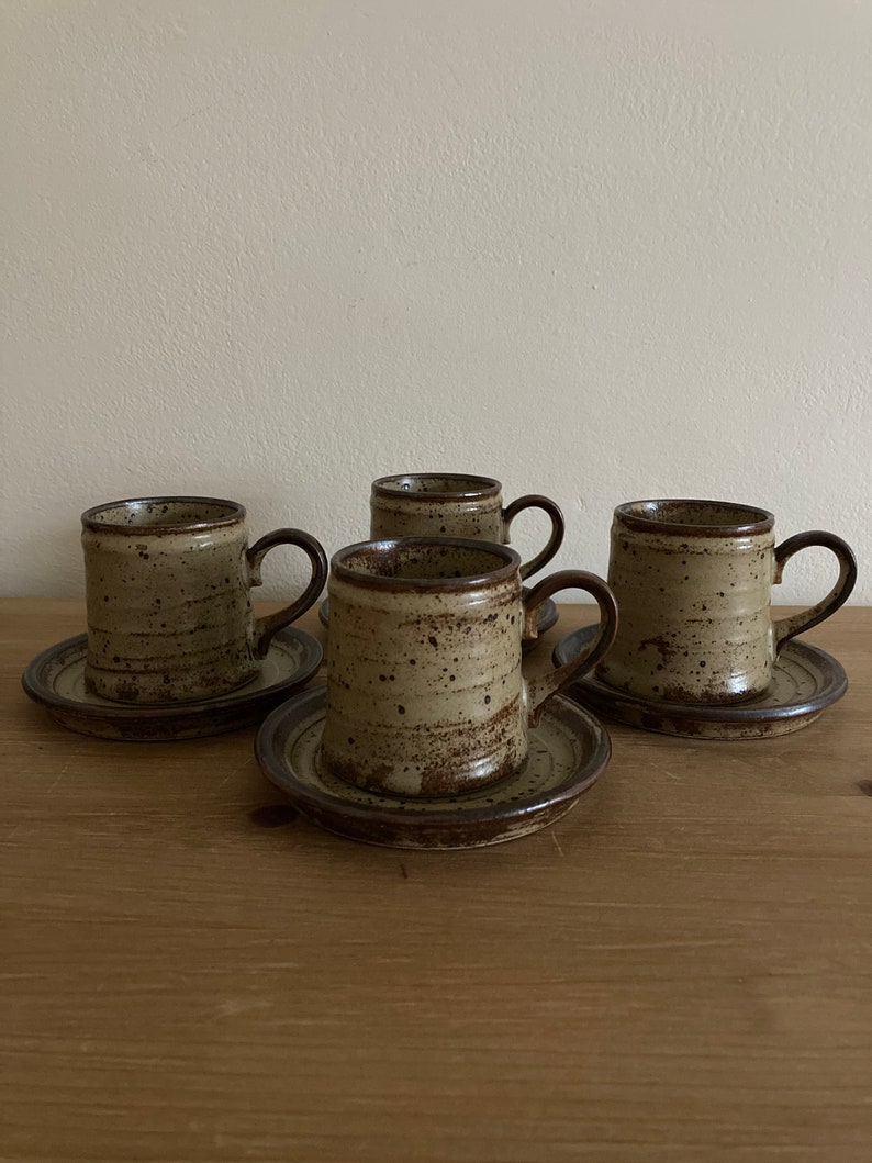 May include: Set of four brown and beige ceramic coffee mugs with matching saucers. The mugs have a speckled glaze and a brown rim.