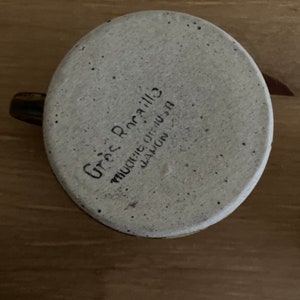 May include: A white ceramic mug with a speckled glaze. The mug has a brown handle and the words "Greg Rocallo" and "Muncie, Indiana" written on the bottom.