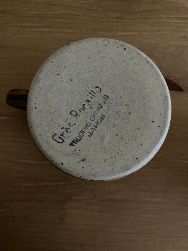 May include: A white ceramic mug with a speckled glaze. The mug has a brown handle and the words "Greg Rocallo" and "Muncie, Indiana" written on the bottom.