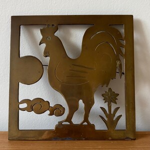 May include: A brass-coloured trivet with a rooster design. The rooster is in profile, with a sun, clouds, and a flower. The trivet has a square frame with cut-out details. The rooster is standing on a small base.
