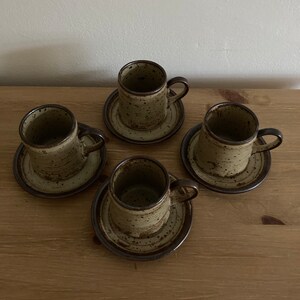 May include: Set of four brown and beige speckled ceramic coffee cups and saucers. The cups have a rounded shape and a small handle. The saucers have a slightly raised rim.