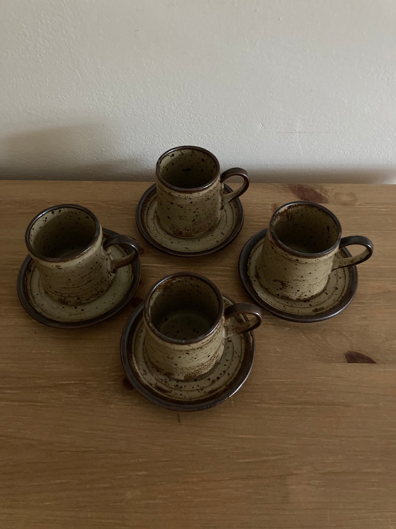 May include: Set of four brown and beige speckled ceramic coffee cups and saucers. The cups have a rounded shape and a small handle. The saucers have a slightly raised rim.