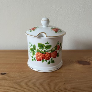 May include: White ceramic sugar bowl with a lid and a gold trim. The bowl is decorated with a pattern of red strawberries and green leaves.
