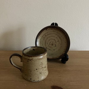 May include: A speckled brown and beige ceramic coffee cup and saucer set. The cup has a handle and the saucer has a circular design.