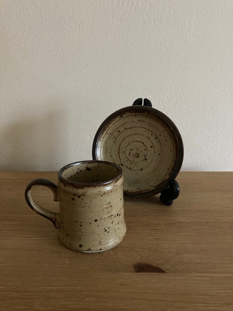 May include: A speckled brown and beige ceramic coffee cup and saucer set. The cup has a handle and the saucer has a circular design.