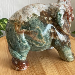 May include: A green, brown and white carved stone elephant figurine. The elephant is standing on a wooden surface.
