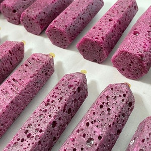 May include: Close-up of several purple, porous, rectangular obelisks with a yellow tip. The obelisks are arranged on a white surface. The objects have a textured appearance and are a deep shade of purple.