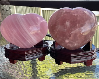 Puede incluir: Dos grandes cristales de cuarzo rosa en forma de corazón sobre soportes de madera. Los cristales están pulidos y tienen una superficie lisa y brillante.