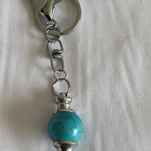 May include: A silver key chain with a turquoise stone pendulum. The pendulum is attached to a silver chain and a silver ring.
