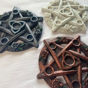 May include: Three pentagram candle holders with a triquetra design in the center. The candle holders are made of a textured material and have a rustic look. The candle holders are in black, white, and copper.