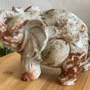 May include: A hand-carved elephant figurine made of white, brown, and green marble. The elephant is standing with its trunk raised and its head tilted to the side.