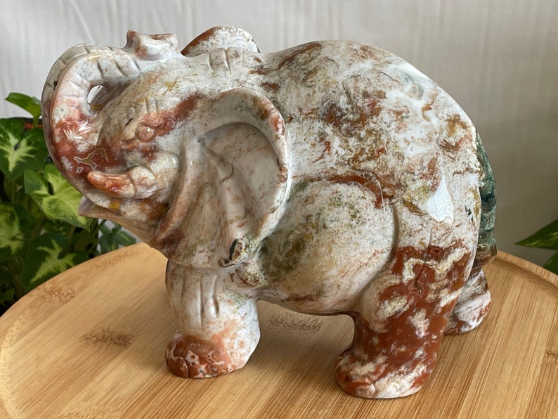 May include: A hand-carved elephant figurine made of white, brown, and green marble. The elephant is standing with its trunk raised and its head tilted to the side.