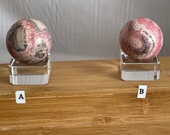 May include: Two pink and gray gemstone spheres on clear stands, labeled A and B. The spheres are on a wooden surface.