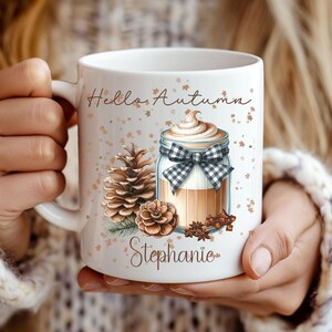 Puede incluir: Taza de cerámica blanca con el texto "Hello Autumn" y el nombre "Stephanie". La taza tiene un tema otoñal con un gráfico de un tarro con crema batida, una piña y especias.