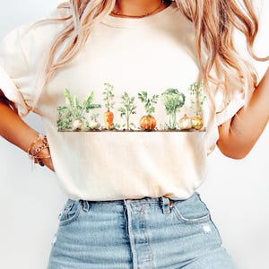 Op de afbeelding: Een wit T-shirt met een aquarelontwerp van groenten, waaronder wortels, broccoli, knoflook en uien. De groenten zijn in een rij over de voorkant van het shirt gerangschikt.
