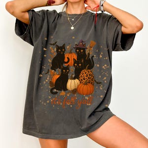 Puede incluir: Camiseta gris oscuro con diseño de Halloween. El gráfico incluye gatos negros, calabazas y el texto "it's fall y'all". Los gatos llevan sombreros de bruja y escobas, rodeados de hojas de otoño.