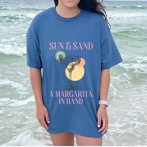 Op de afbeelding: Een blauw t-shirt met de tekst "Sun & Sand, A Margarita in Hand" en een afbeelding van een margarita-glas met limoenpartjes.