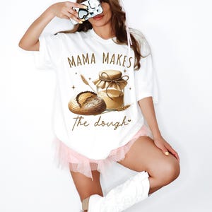 Può includere: Maglietta bianca con una grafica marrone di un barattolo di pasta e una pagnotta di pane. Il testo "Mama Makes the Dough" è stampato sulla maglietta.