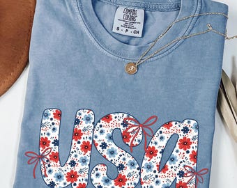 May include: A blue Comfort Colors t-shirt with the word "USA" in a floral pattern of red and blue flowers. The shirt has a small tag and is folded. A gold necklace with a pendant is draped across the shirt.
