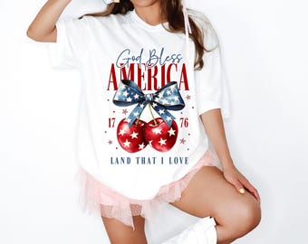 May include: White t-shirt with a patriotic design. The shirt features the text "God Bless America" in red and blue, a blue bow, and two cherries with star patterns. The text "Land That I Love" is below the cherries.