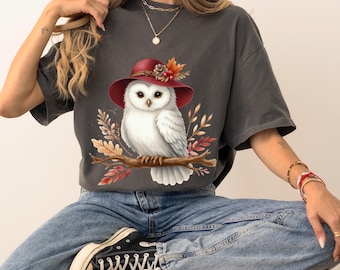 May include: Dark grey t-shirt featuring a white owl wearing a red hat adorned with fall foliage. The owl is perched on a branch, surrounded by autumn leaves. The shirt is paired with blue jeans and black sneakers.
