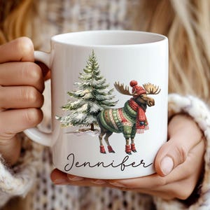 May include: White ceramic mug featuring a festive moose wearing a red hat, scarf, and sweater, standing next to a snow-covered Christmas tree. The name "Jennifer" is written in cursive script below the moose.