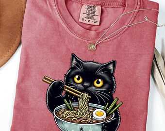 May include: A red t-shirt with a black cat eating ramen noodles from a bowl. The cat is holding chopsticks and the bowl has Japanese characters on it. The text on the tag says "Comfort Colors".