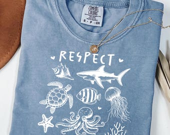 May include: A light blue t-shirt with a white graphic design that says "Respect the Locals" and features a variety of sea creatures including a shark, a turtle, an octopus, a starfish, a fish, and a jellyfish.