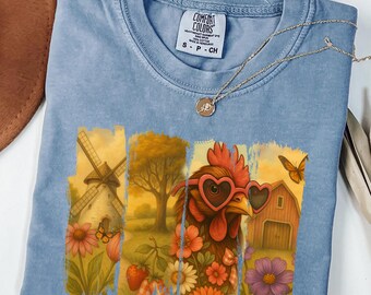 May include: A light blue t-shirt with a graphic print of a rooster wearing heart-shaped sunglasses. The rooster is surrounded by flowers and a windmill in the background. The t-shirt has a tag that says "Comfort Colors" and "S-P-CH".