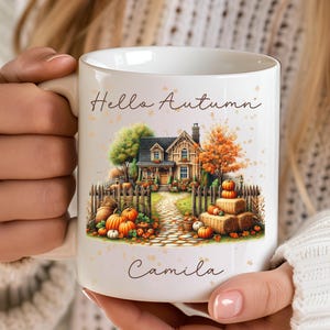 Op de afbeelding: Witte keramische mok met een herfstthema. De mok heeft een gedetailleerde illustratie van een huis, pompoenen en de tekst "Hello Autumn" en "Camila". Het ontwerp bevat oranje, groene en bruine kleuren.