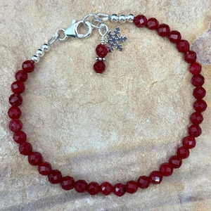 May include: A bracelet composed of small, faceted deep red beads strung together. The piece features a silver-toned lobster claw clasp and a dangling charm shaped like a snowflake, accented by a single matching red bead on the charm attachment.