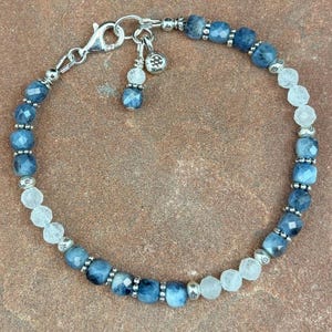 May include: A beaded bracelet featuring alternating faceted blue and clear gemstones. The bracelet is strung on a silver chain with a lobster clasp closure. Small silver beads separate the larger gemstones.