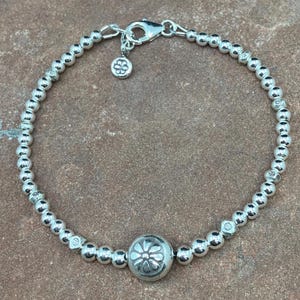 May include: A silver beaded bracelet with a floral charm and a larger floral-detailed bead. The bracelet features small, round beads strung together, with a lobster clasp closure. The charm and the larger bead have a flower design.