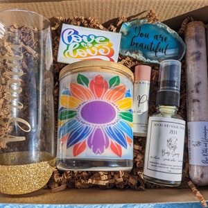 May include: A gift box filled with self-care items, including a rainbow candle, a glass with glitter, a bottle of body spray, a tube of bath salts, a sticker that says "Love is Love", and a stone that says "You are beautiful".